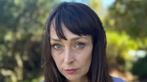 Juliette Bryant, now aged 43, has long brown hair with a fringe and stands outside with a serious expression 